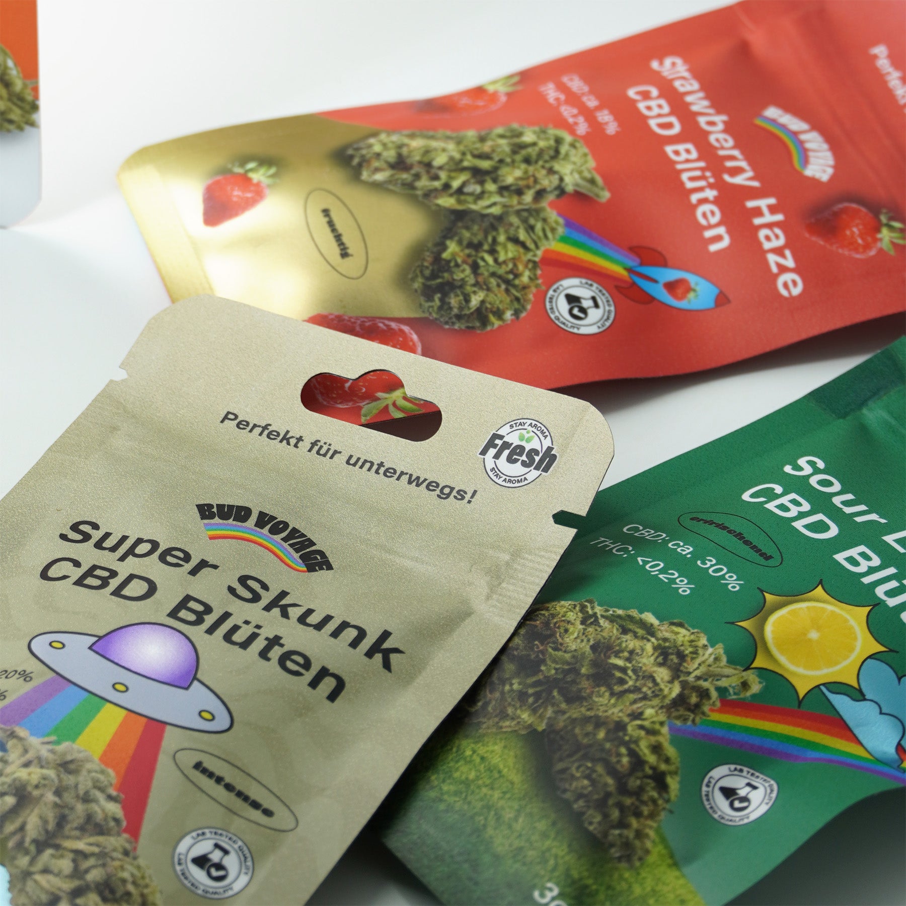 Fotografie produktu: Tři balíčky CBD květů zobrazují Super Skunk s UFO a duhou, Strawberry Haze s jahodami a Sour Lemon s citronem, všechny s pečetěmi z laboratorních testů a barevné.

