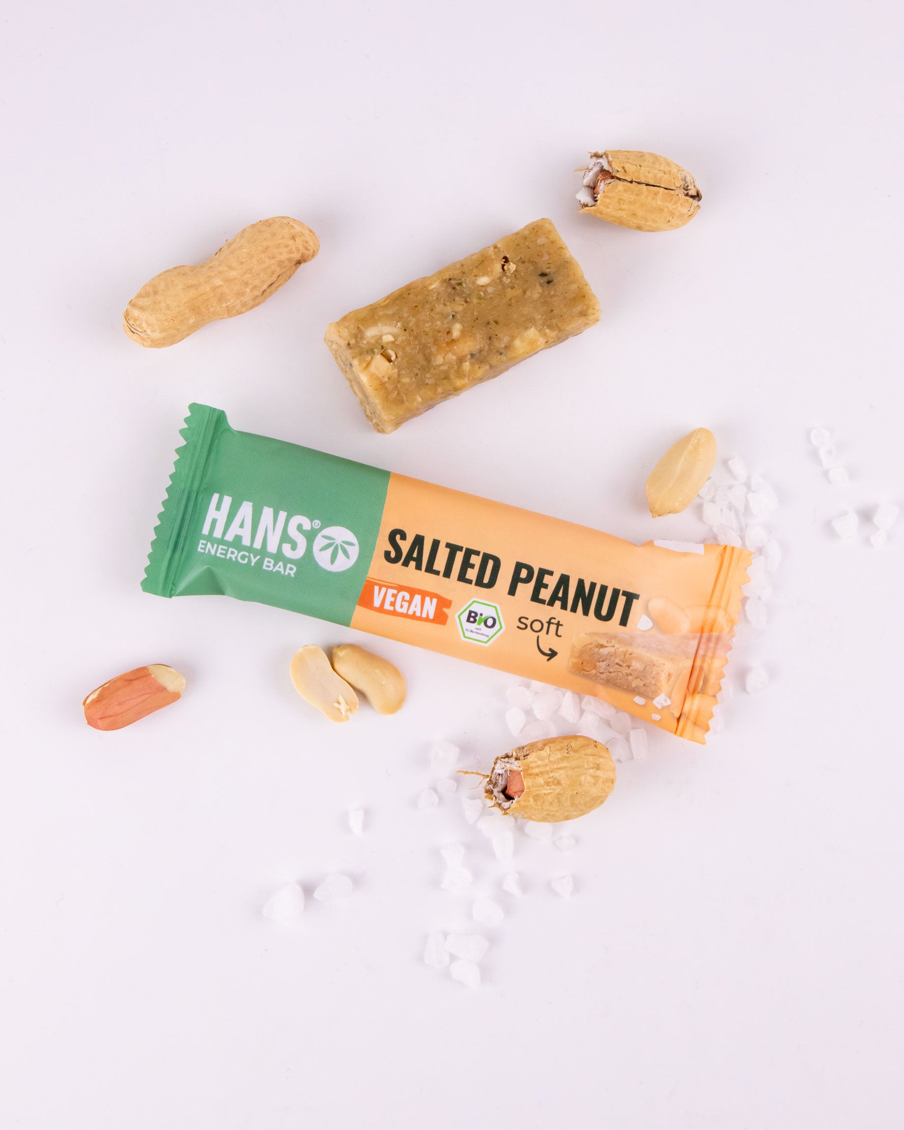 Snímek produktu Hans Salted Peanut Vegan Energy Bar na bílém pozadí s arašídy a krystalky mořské soli zobrazující měkkou, veganskou, bio tyčinku se zeleným a oranžovým obalem.