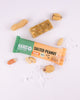 Produktový snímek veganské energetické tyčinky Hans Salted Peanut na bílém pozadí s arašídy a krystalky mořské soli, který ukazuje design obalu a jemnou texturu tyčinky.
