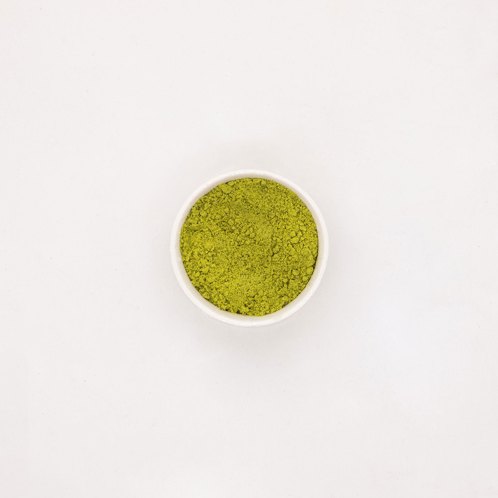 Fotografie: Eine Schale mit hellgrünem, körnigem Matcha-Pulver auf weißem Hintergrund, das Pulver hat eine leicht unebene Textur und eine satte Farbe
