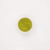 Fotografie: Eine Schale mit hellgrünem, körnigem Matcha-Pulver auf weißem Hintergrund, das Pulver hat eine leicht unebene Textur und eine satte Farbe
