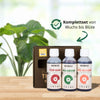 Product shot of Biobizz Top Max, Bio Grow, and Bio Bloom plant nutrients on a light wood surface against a blurred background of lush green leaves, showcasing the dark brown bottles, vibrant red, green, and orange label accents, and a brown cardboard box
