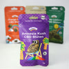 Bud Voyage product packaging, Amnesia Kush CBD flowers, showing a smiling cannabis flower on a deckchair in front of palm trees and a rainbow, purple packaging with checkerboard pattern and quality seal

