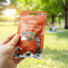 Bud Voyage product photography: A hand holds an orange-colored pack of Orange COW CBD flowers outdoors against a blurred background with trees and a rainbow on the packaging, which shows green flowers and a cute cow motif
