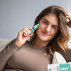 Photography of a woman holding a small, teal-colored bottle of 10% HA serum against her face while sitting on a beige couch wearing a mauve top and gold necklace