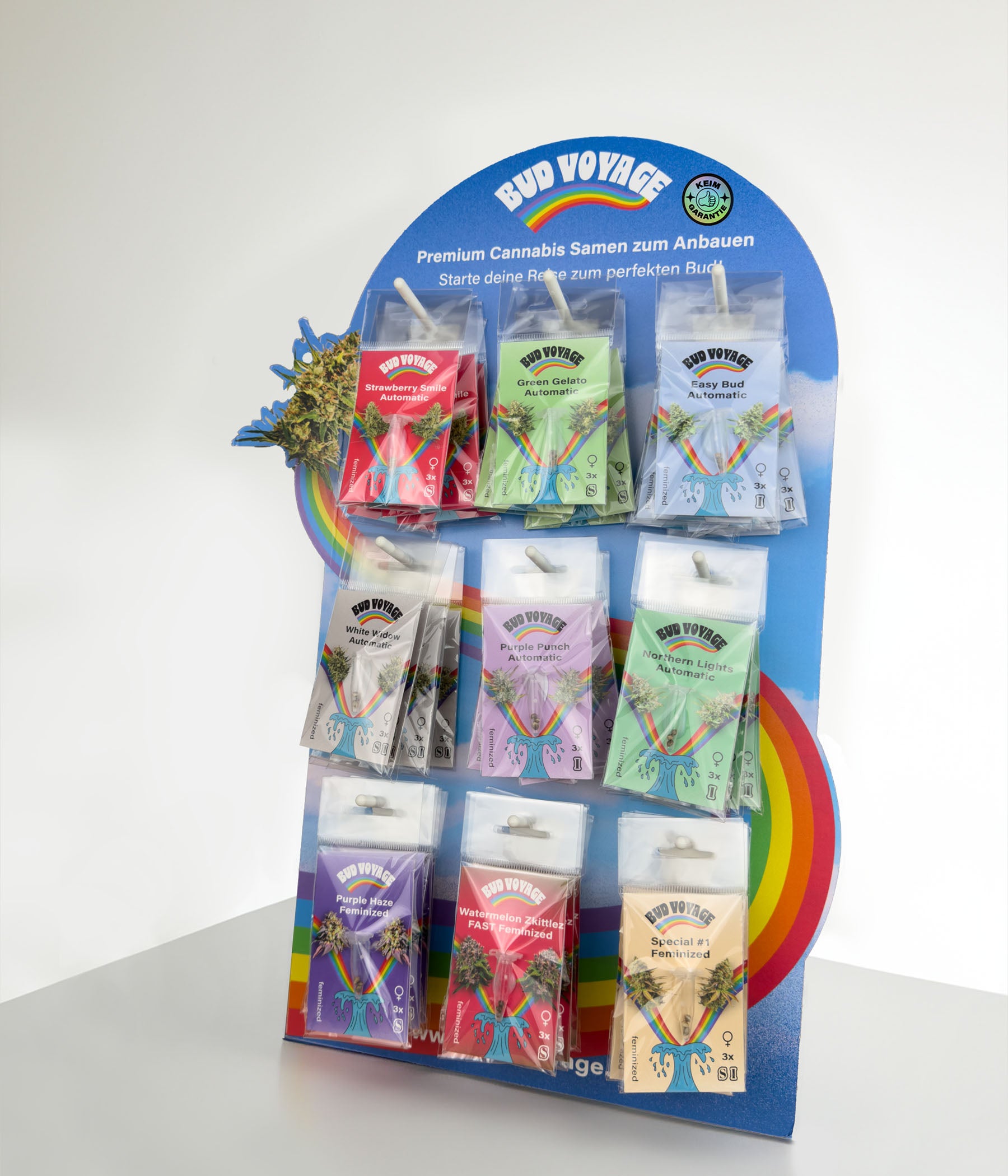 Product photo: Bud Voyage cannabis seed display with different strains such as Strawberry Smile, Green Gelato and Northern Lights, presented on a rainbow-colored stand with wave and cannabis leaf motifs in bright colors.
