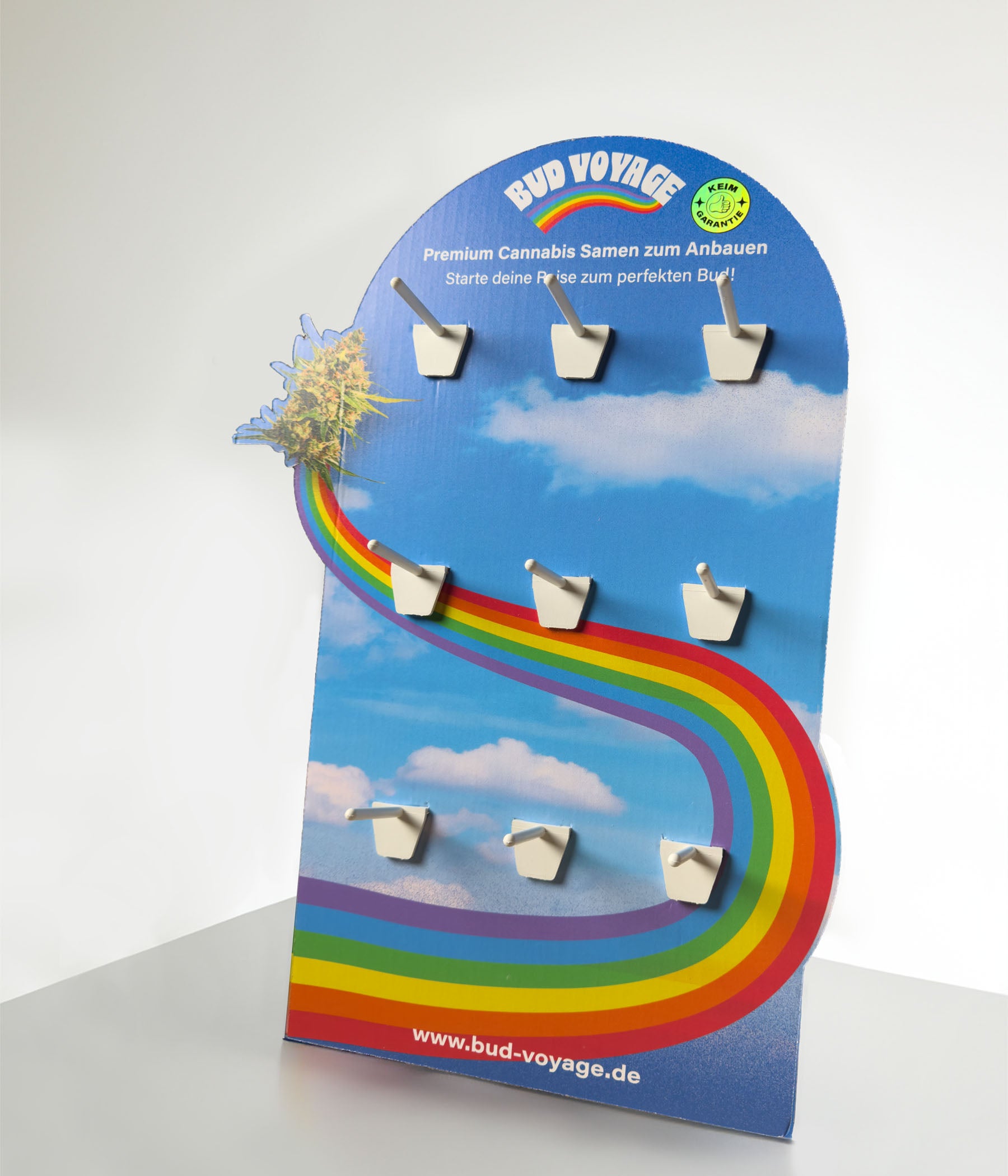 Kartonaufsteller von Bud Voyage zeigt Cannabis-Samen-Display vor einem blauen Himmel mit Regenbogen und weißen Haltern, die Marihuana-Blüten und die Webseite wwwbud-voyagede darstellen

