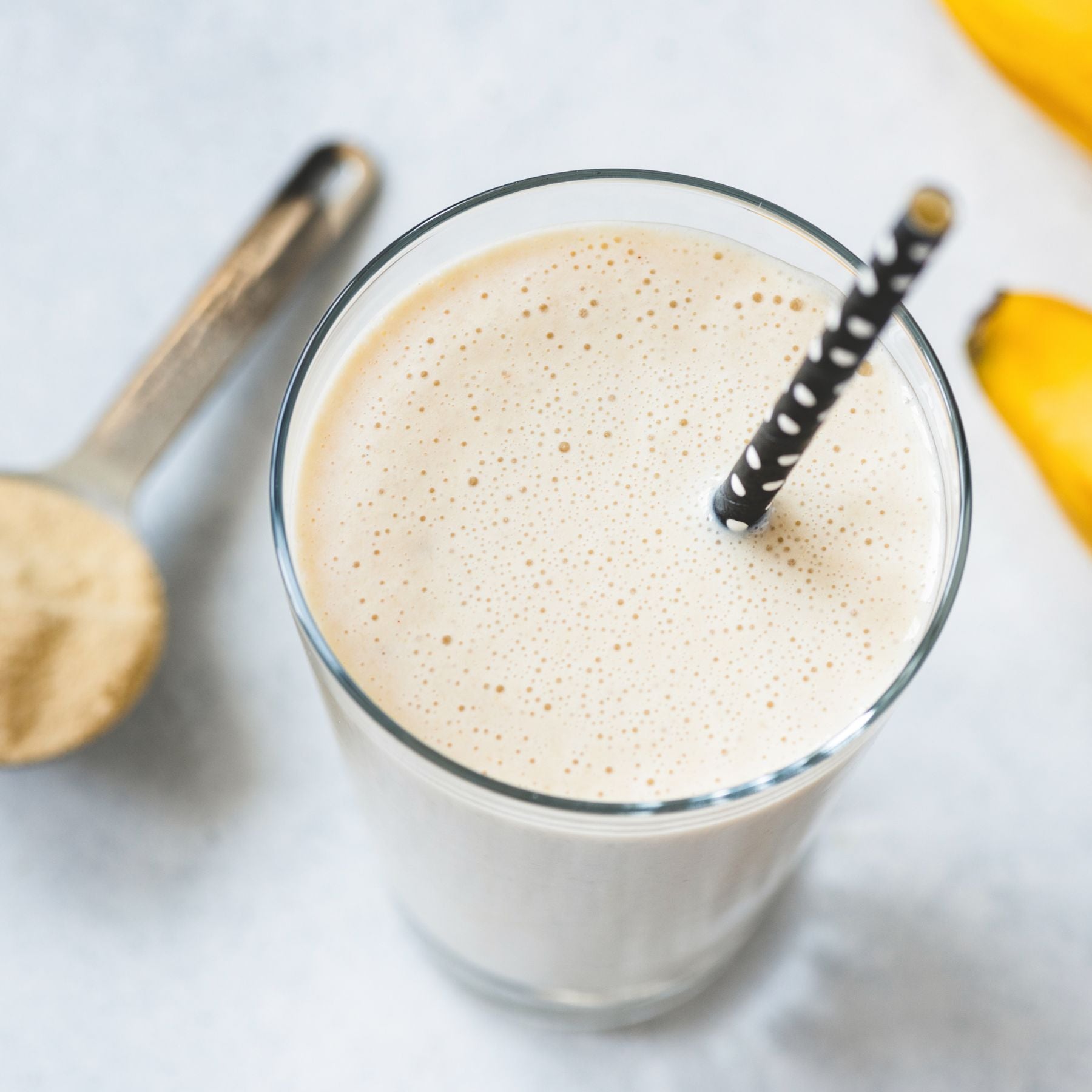 Photo: A creamy banana smoothie in a glass with a black straw next to a measuring spoon with light powder and bananas shown on a white background, with a fine-pored texture and creamy color