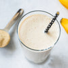 Photo: A creamy banana smoothie in a glass with a black straw next to a measuring spoon with light powder and bananas shown on a white background, with a fine-pored texture and creamy color