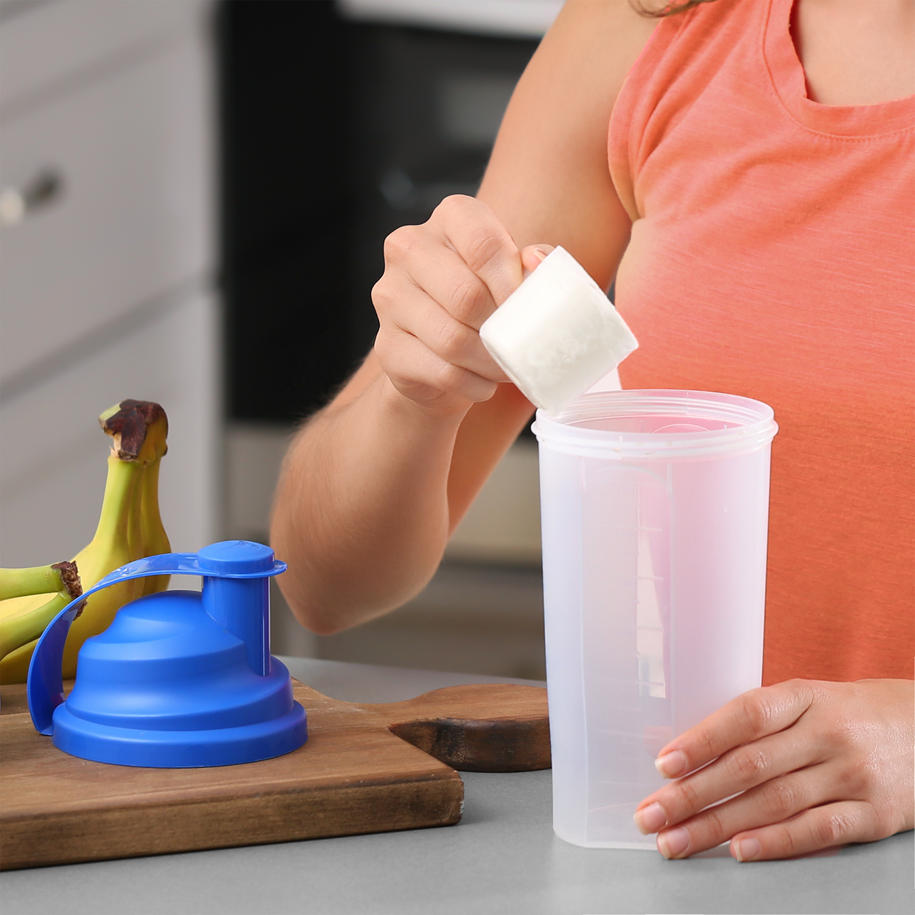 Photograph Woman adding protein powder to shaker cup in kitchen with vibrant blue lid and ripe yellow bananas a wooden cutting board is nearby
