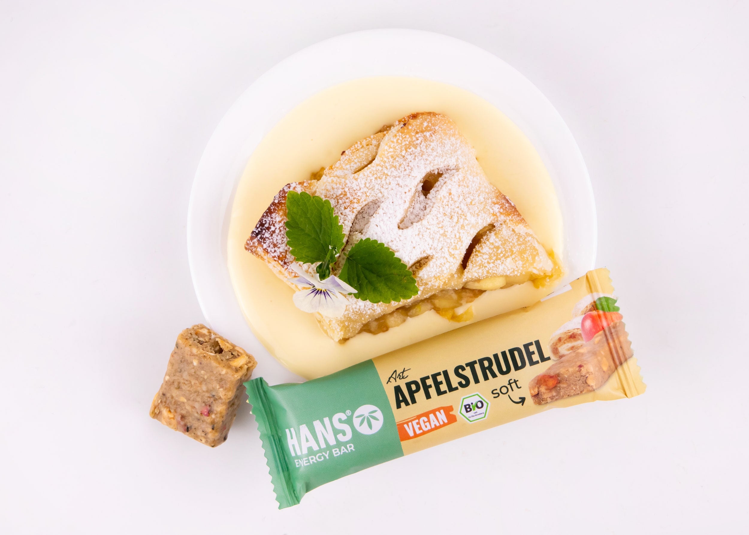 Food photography showing a slice of apple strudel with vanilla sauce garnished with mint and a pansy on a white plate next to a vegan apple strudel energy bar featuring vibrant colors and a dusting of powdered sugar
