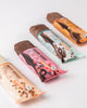 Produktfotografie unbekannter Künstlerin zeigt drei Bio- und vegane Schokoriegel auf weißem Hintergrund mit Kokos, Himbeeren und Nüssen in zarter rosa, mint und beige Verpackung.
