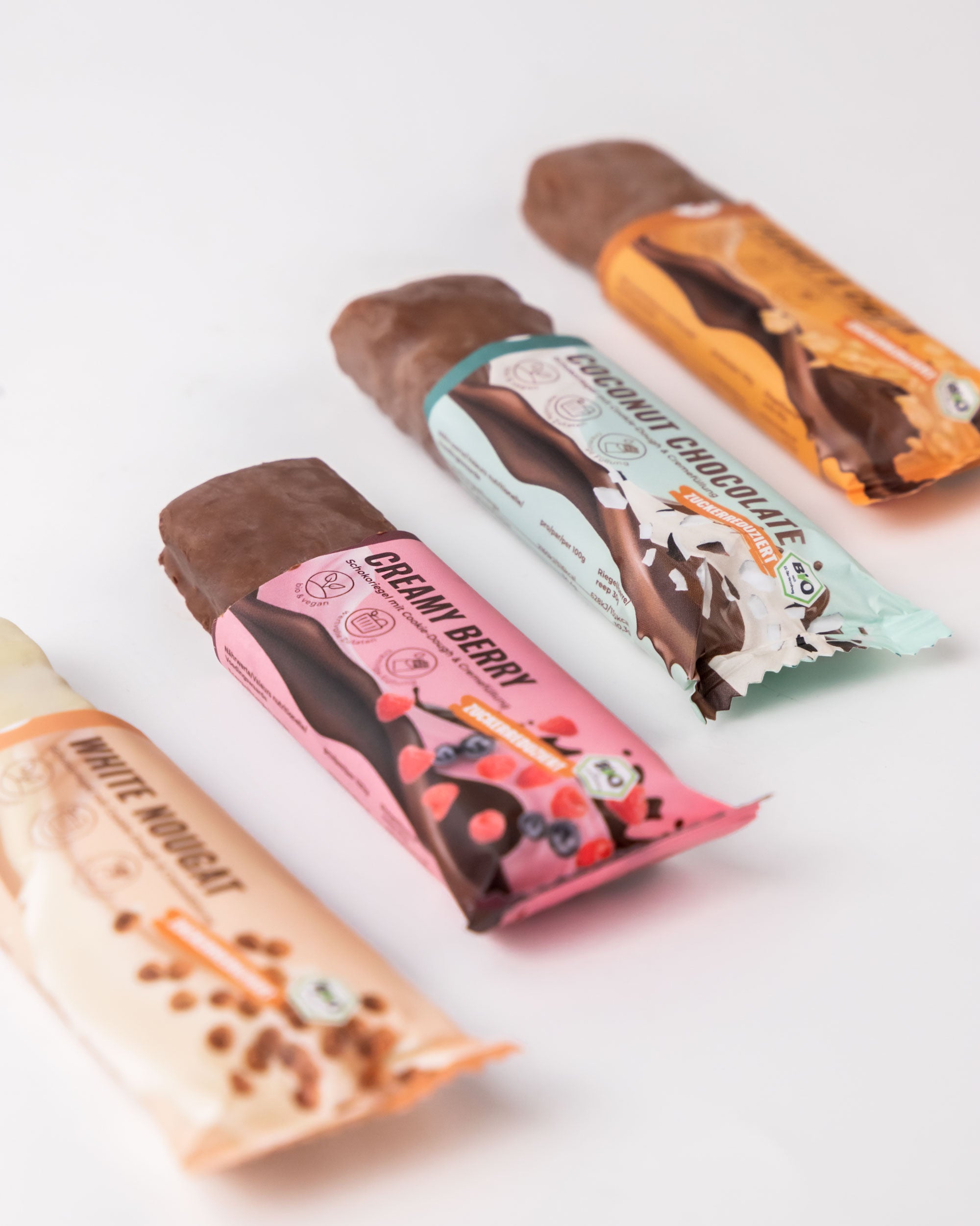 Produktfotografie von drei Bio- und veganen Schokoriegeln auf weißem Hintergrund mit Kokos, Himbeeren und Nüssen in den jeweiligen Verpackungen
